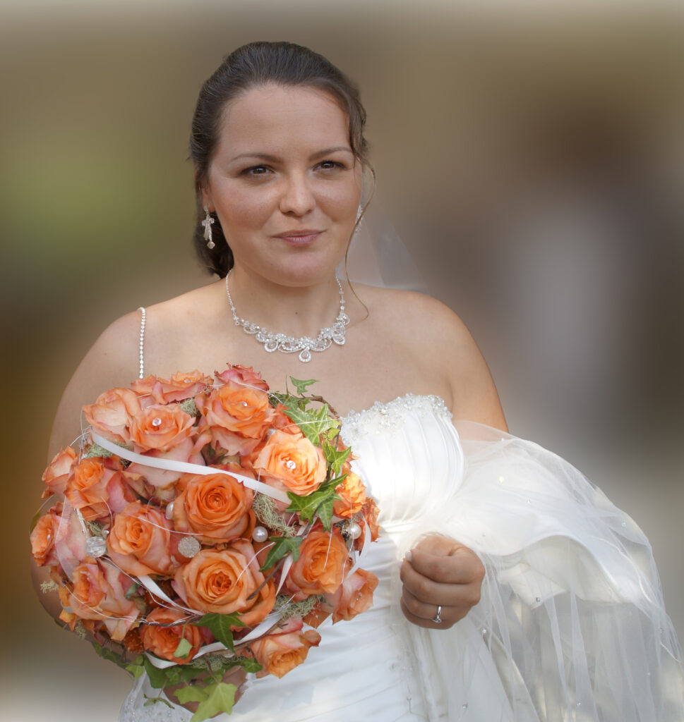 Eine Braut mit wunderschönen Diamantenen Ohrringen und Collier schaut lächelnd in die Kamera. Sie hält in der rechten Hand einen Brautstrauss aus Orangefarbenen Rosen – mit der rechten Hans hält sie ihren Schleier.