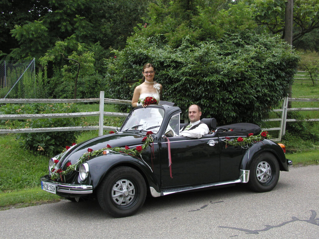 Ein hochzeitlich  mit roten Rosen geschmücktes schwarzes VW Käfer Cabrio steht auf einer alten Landstraße vor einer grünen Landschaft mit Holzzaun. Im Wageninneren sitzt der Bräutigam lässig, den Arm auf der Autotüre aufgestützt, am Steuer. Die Braut steht im Beifahrerraum lächelt und hat ihren Brautstrauß in der Hand