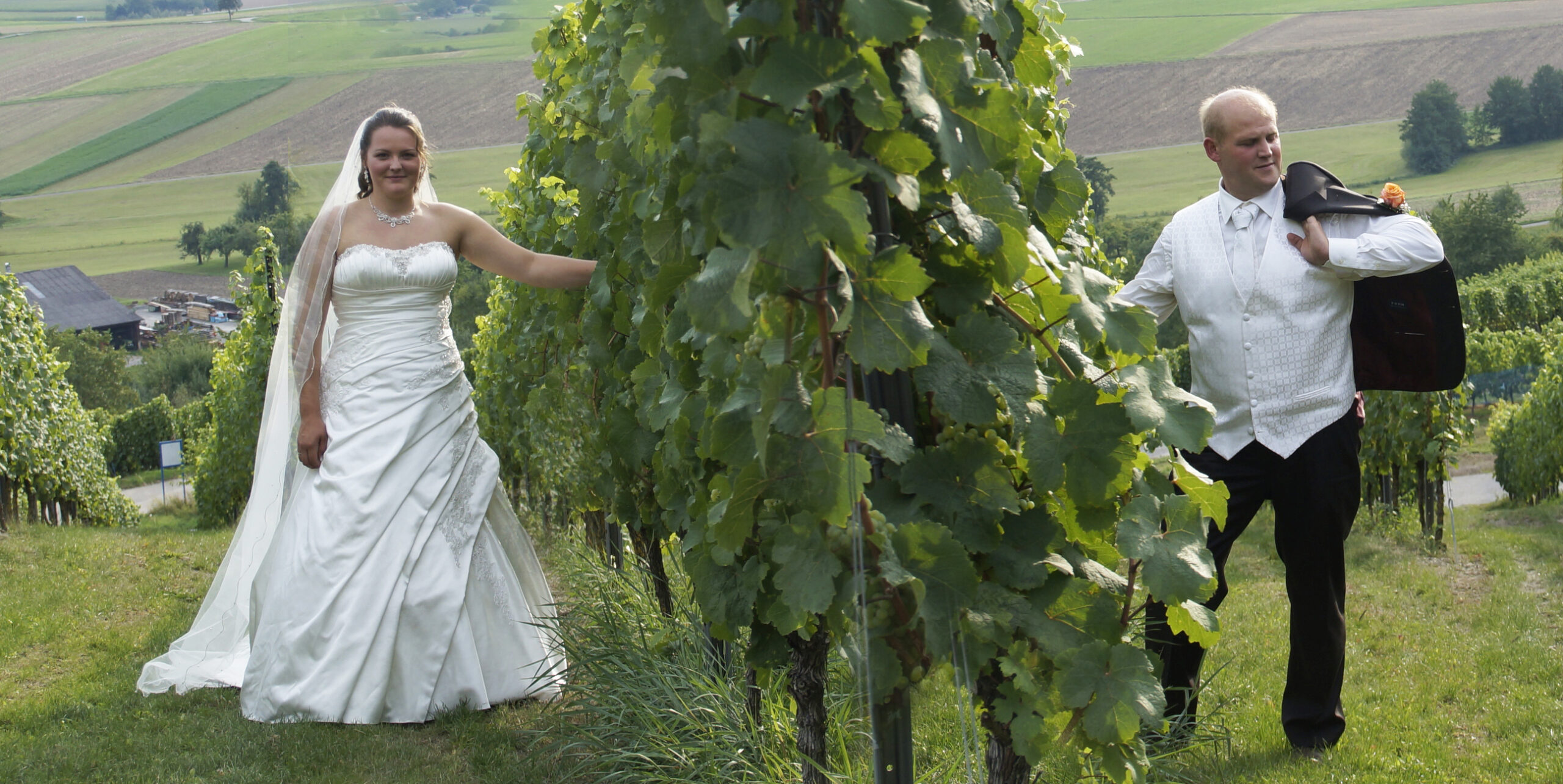 Braut und Bräutigam laufen rechts und links von Weinreben.
Der Bräutigam trägt sein dunkles Jacket über der linken Schulter. Weisses Hemd und Weste schaffen die Verbindung zum Hochzeitskleid der Braut. Da die Reihe der Rebstöcke vorne zuende ist assoziiert man ein Treffen des Brautpaares in Kürze