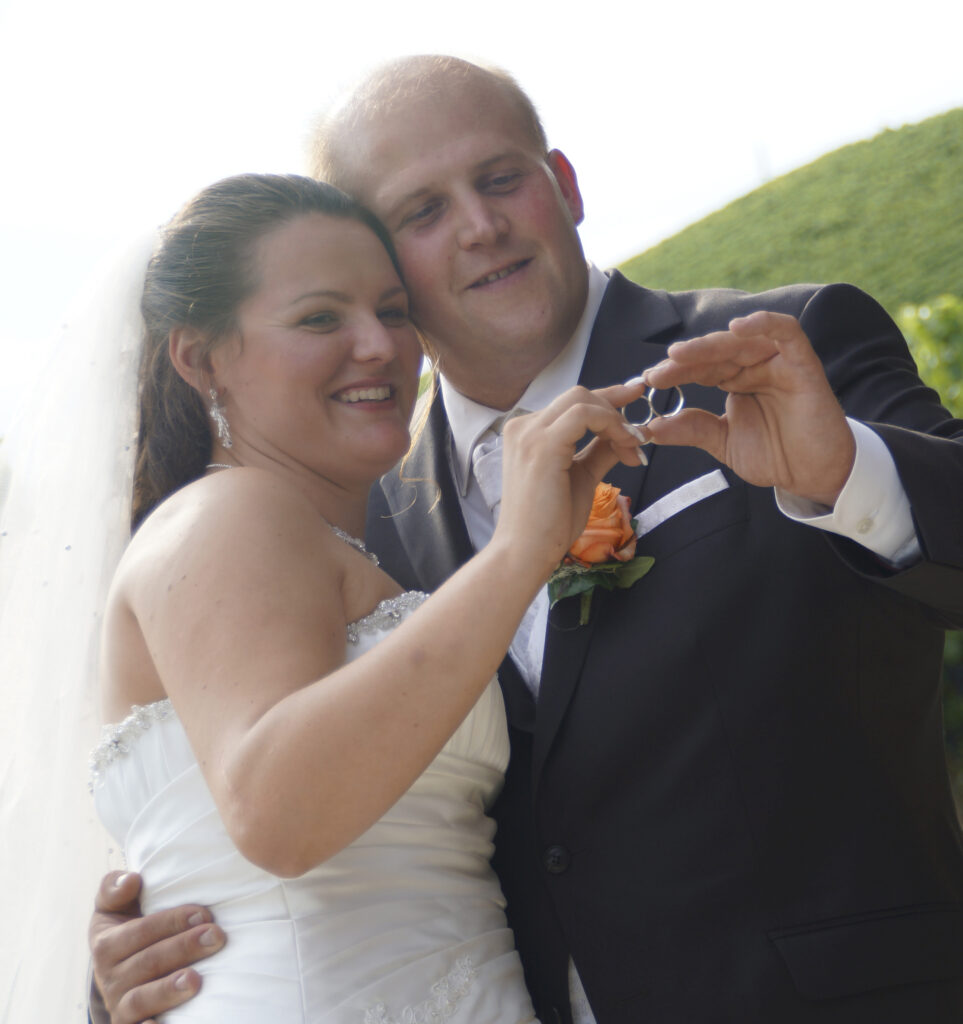 Ein sich umarmendes Brautpaar hält die Ringe aneinander und schauen lächeld durch die Ringe hindurch.