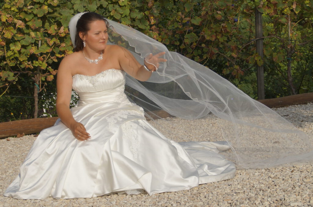 Eine Braut im weissen Brautkleid sitzt auf hellem Kiesboden, das Kleid ist weit ausgebreitet, Der Schleier weht im Wind. Ihre rechte Hand ist erhoben um den Schleier festzuhalten. Licht und Schatten lassen das Brautkleid weich und seidig glänzen.