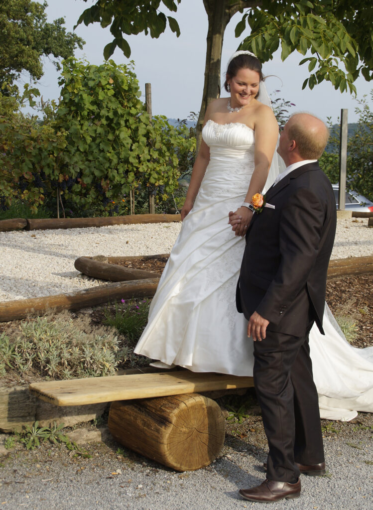 Eine glückliche lächelnde Braut im weißen Brautkleid steht auf einer Holzbank und schaut auf ihren Bräutigam herunter. Er steht vor der Bank und schaut zu ihr hinauf. Sie fassen sich an den Händen.
