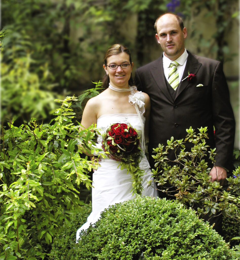 Ein Brautpaar steht in einer wunderschönen Parkanlage.
Der aus roten Rosen bestehende Hochzeitsstrauß den die Braut vor sich trägt, leuchtet vor dem Brautkleid