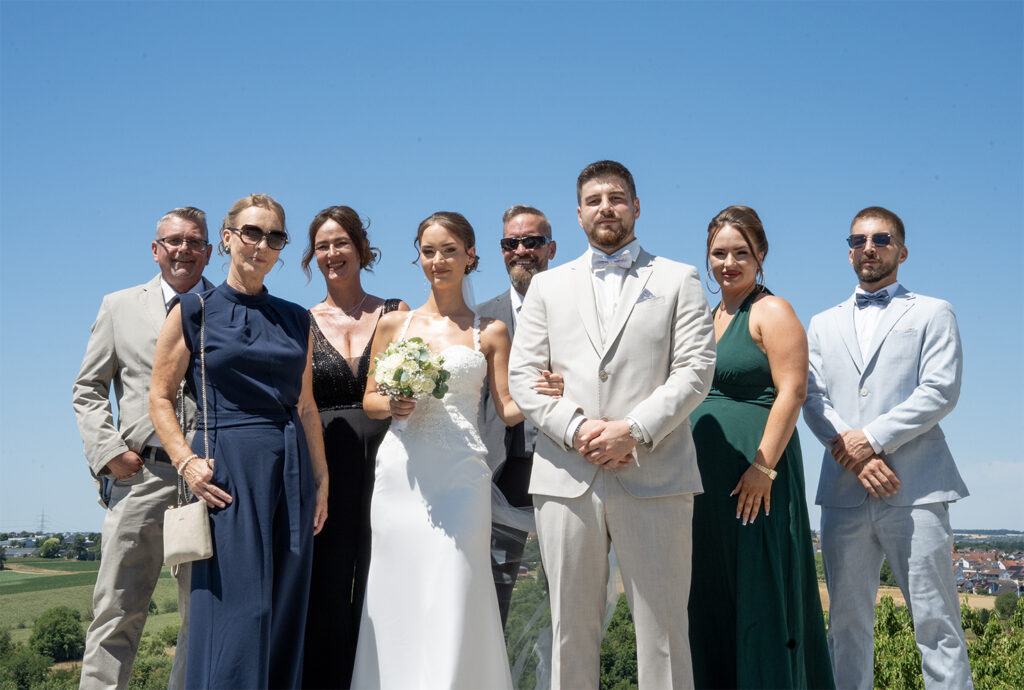 Klassisches Gruppenbild: Das Brautpaar und die Familienmitglieder  stehen in zwei Reihen nebeneinander, alle schauen in die Kamera. Im Hitergund sieht man weit in die Ferne auf die ländliche Umgebung von Marbach, der Himmel ist wolkenlos und strahlend blau.
