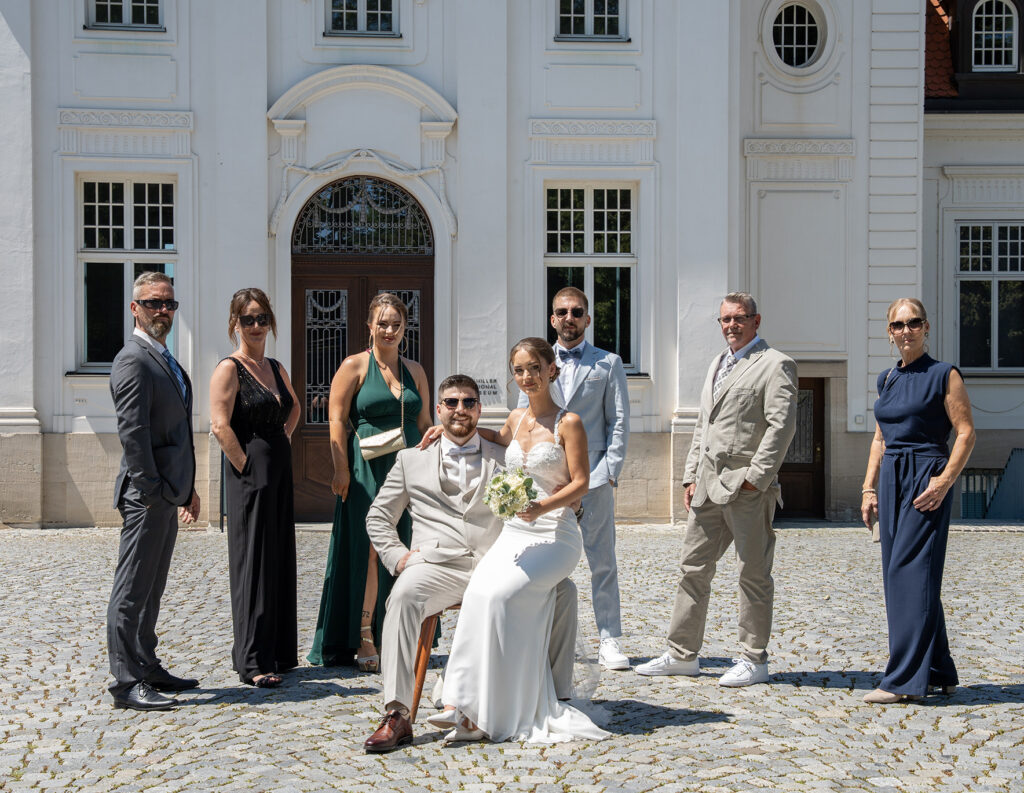 Gruppenbild: Die Familienmitglieder und das Brautpaar vor dem Schiller Nationalmuseum. Die familienmitglieder stehen im Halbkreis, der Bräutigam sitzt auf einem Stuhl und die Braut sitzt auf seinem Schoß.