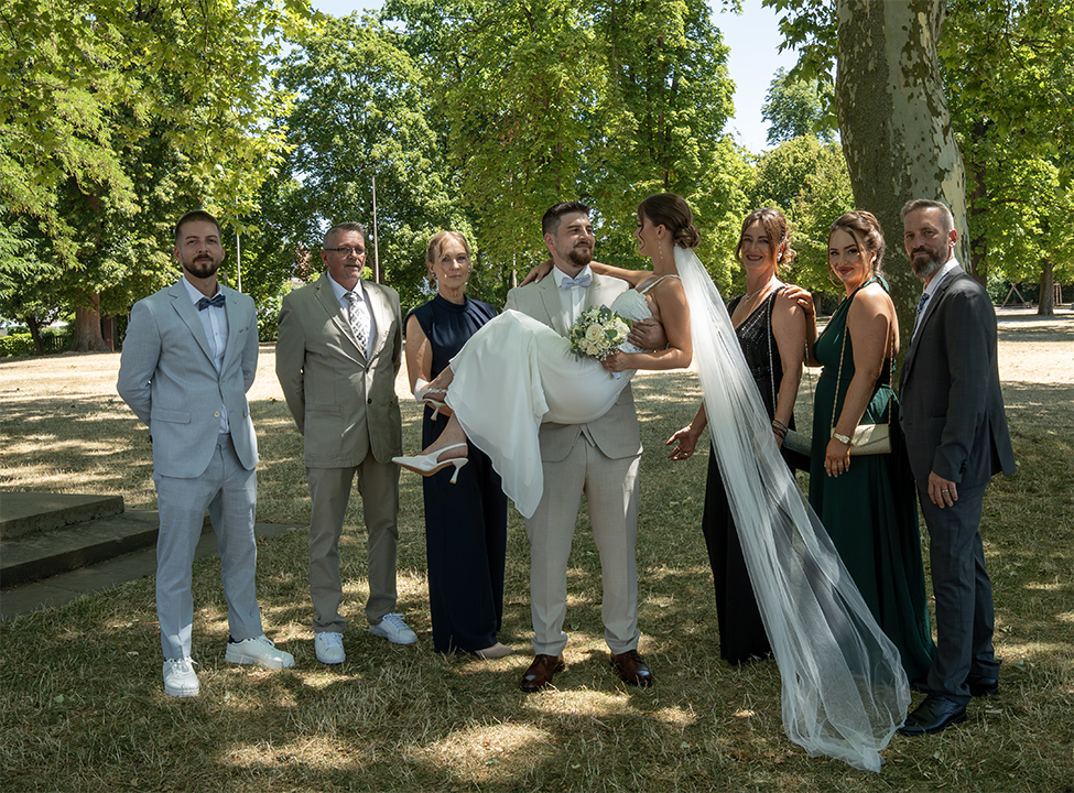 Gruppenbild im mit Platanen bestückten Park der Schillerhöhe: Der Bräutigam trägt die Braut auf seinen Armen, die sechs Familienmitglieder stehen rechts und links des Brautpaars im Halbkreis. Das Brautpaar sieht sich verliebt an, der Schleyer der Braut weht im Wind.
